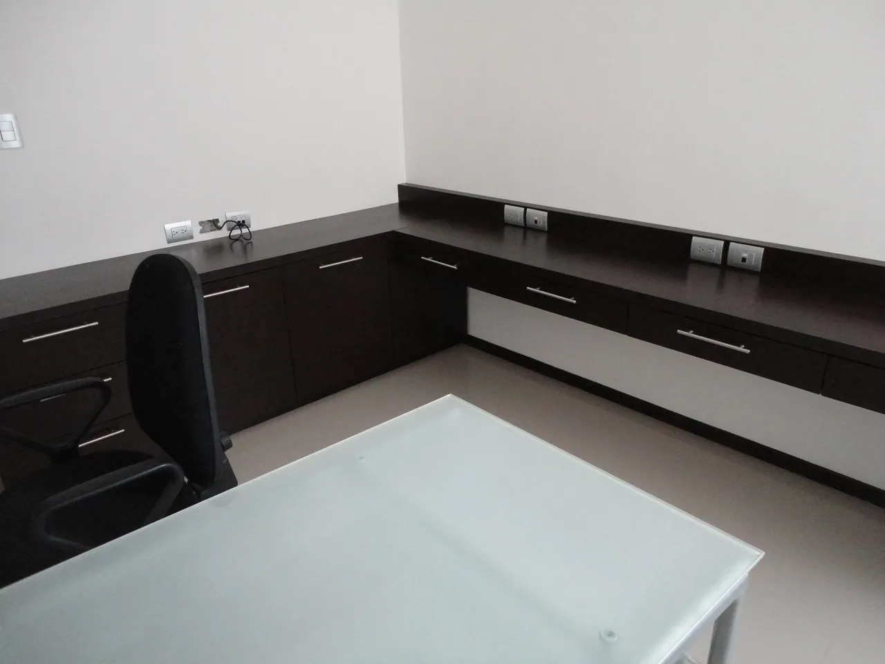 Oficina en casa con barra de trabajo en escuadra, cajonería en madera oscura y cubierta de cristal.