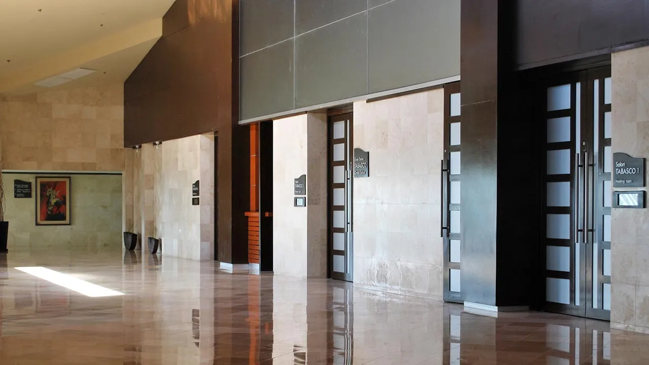 Vestíbulo con puertas altas de madera y plafón oscuro, sobre piso reflejante.