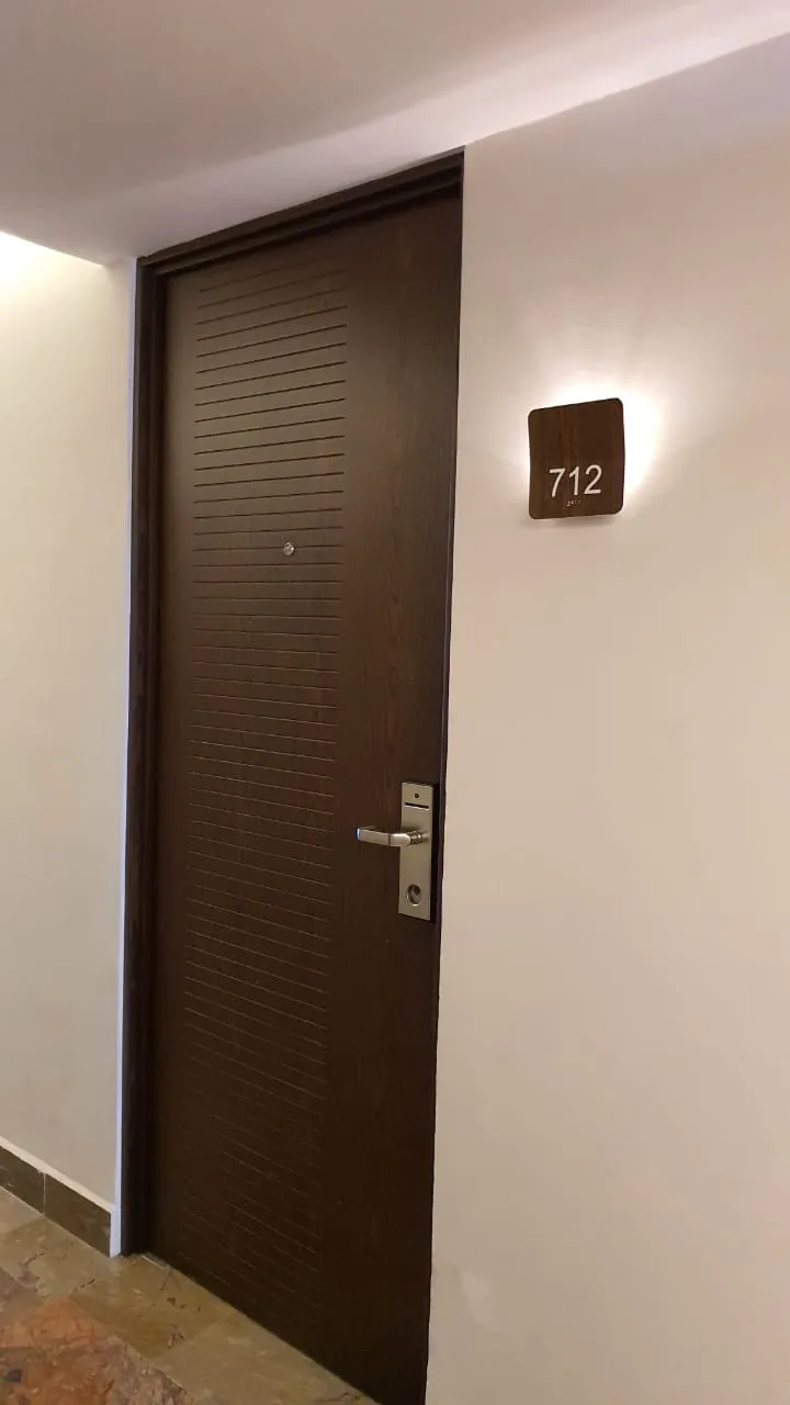 Puerta de habitación en madera oscura con el número de cuarto y aplique de pared.
